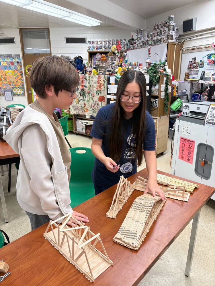 A middle school student helping a younger student with their popsicle-stick bridge design