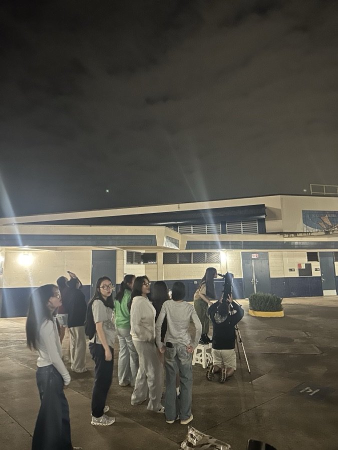 A student looking through a telescope at night while classmates gather around
